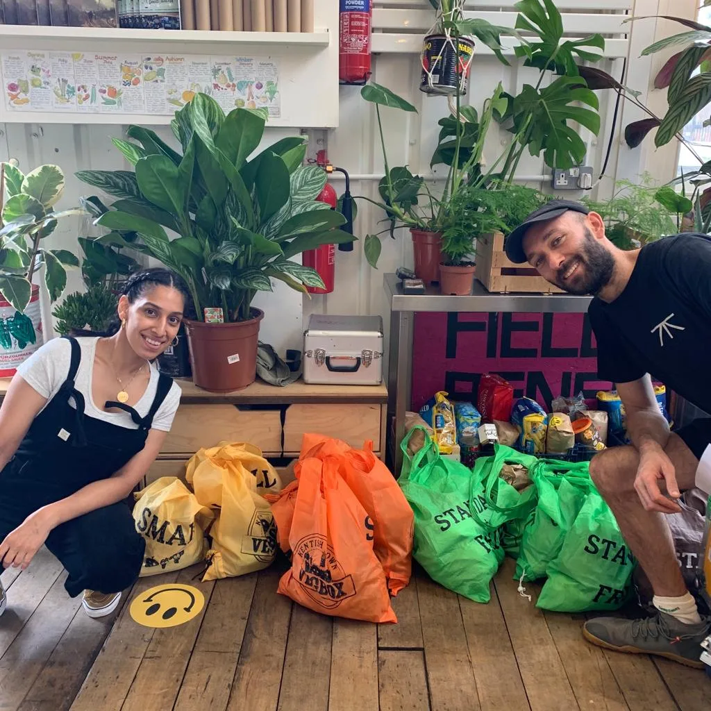 Two smiling volunteers crouched beside a row of colourful Vegbox bags at a collection point