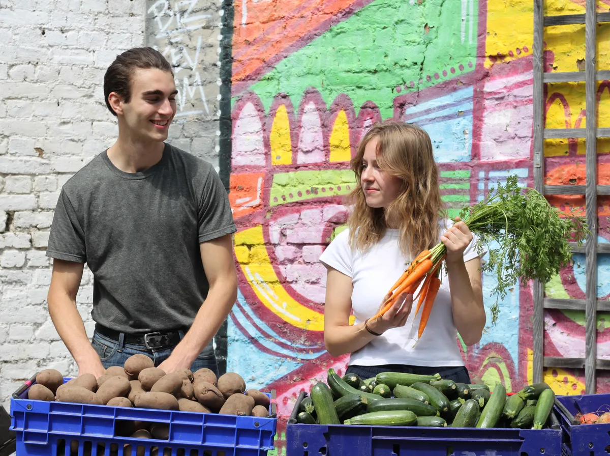 Two Vegbox members in the packing yard, one holding a bunch of carrots, in front of a colourful mural