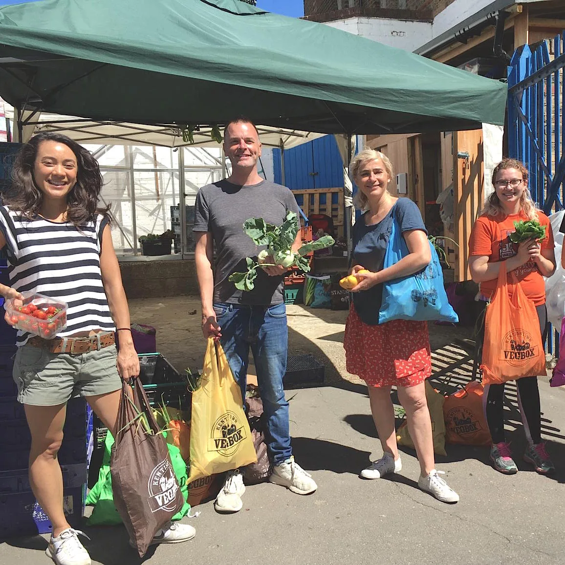 Four smiling Vegbox members at a stall, holding strawberries, kohlrabi and Vegbox bags in the sunshine